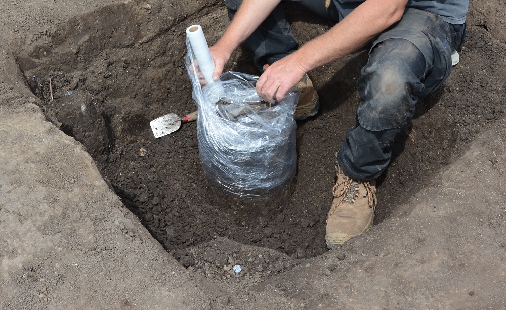 Archäologe hockt im Grabungsschnitt und verpackt im Erdblock einen Bodenfund in Folie.