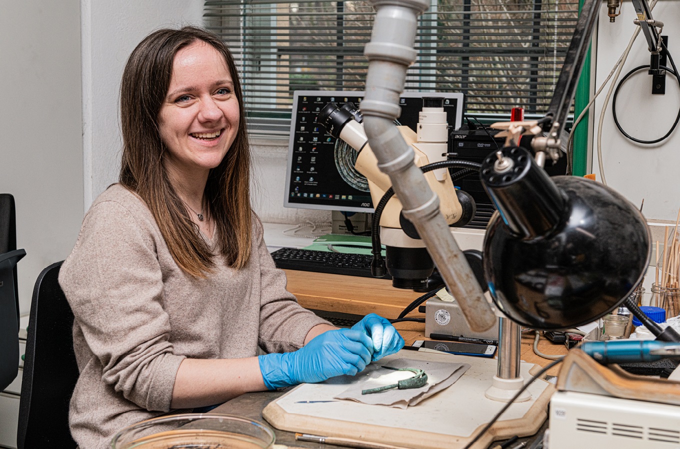 Junge Frau sitzt am Mikroskop in der Restaurierungswerkstatt und legt einen archäologischen Fund frei