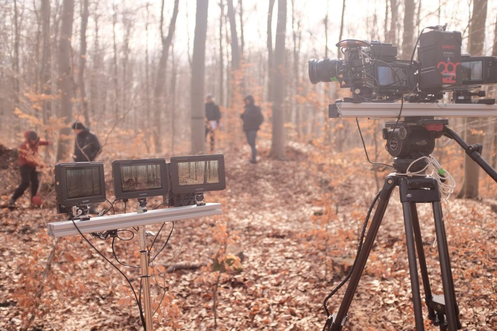 Im Hintergrund herbstlicher lichter Wald mit vier Personen, den Hauptdarstellern des Flmes,, im Vordergrund Filmkamera auf Stativ, mit der Aufnahmen für den Chrononauten erstellt werden.